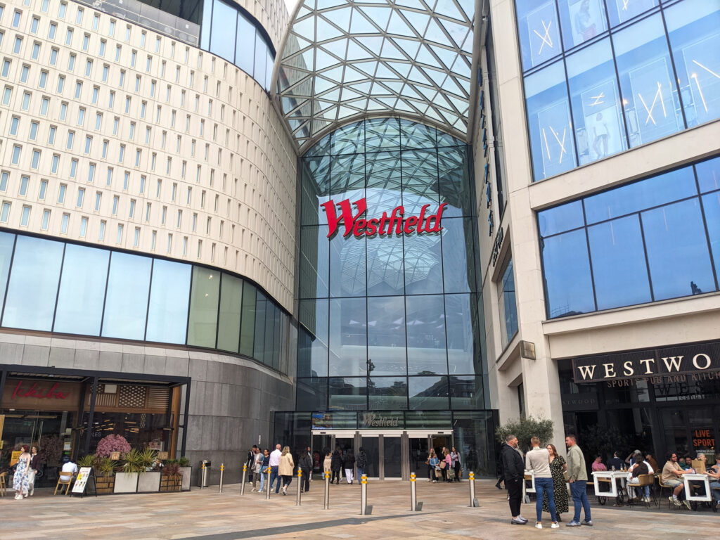 Folk går inn og ut av den store, moderne glassfasaden til kjøpesenteret Westfield London, med restauranter langs siden i et urbant miljø.
