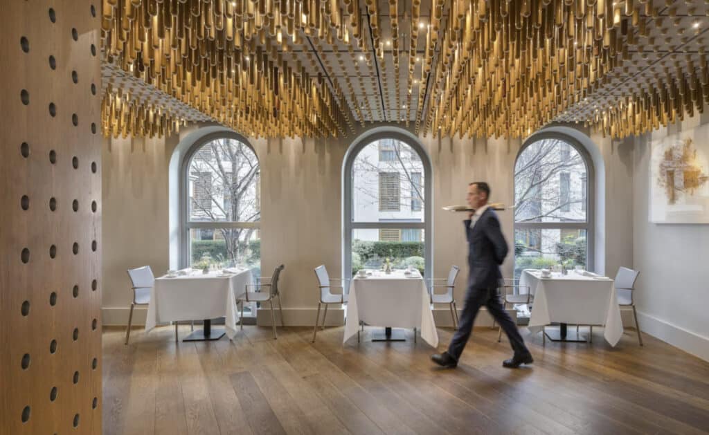 Hotellrestaurant med servitør som bærer noe. Hvite duk på bordene, plassert ved vinduene.