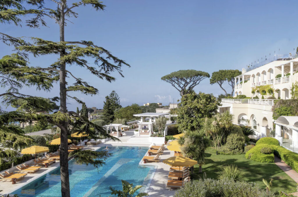 Hotellbasseng med gule parasoller, grønne trær, hvitkalket hotell.