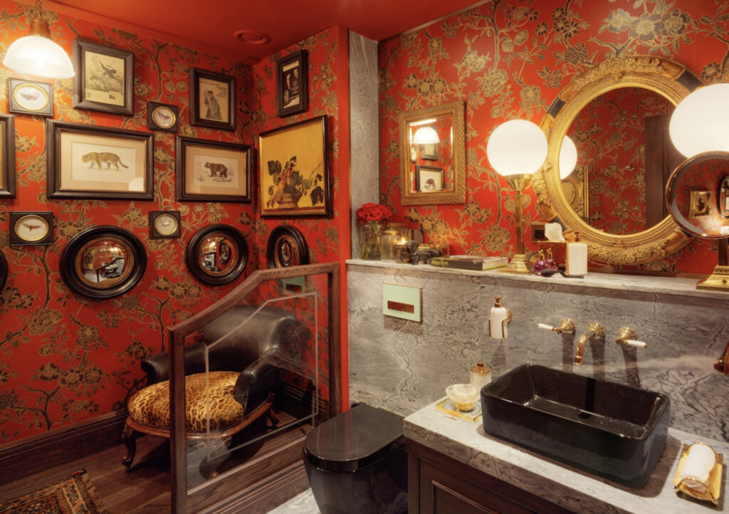 Baderom på rommet med tigermønster på stol, rødt tapet med blomster, bilder på veggene og speil. 