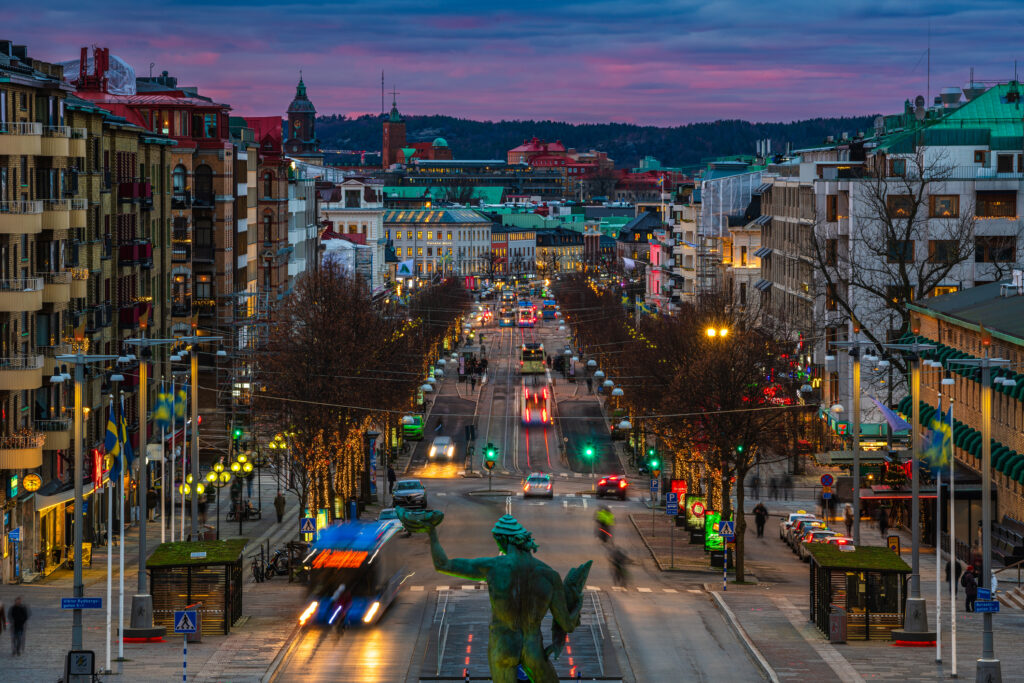Et livlig syn av Avenyn i Gøteborg idet solen går ned, med travle gater, opplyste bygninger og en fargerik himmel. Folk rusler langs fortauene og nyter stemningen.