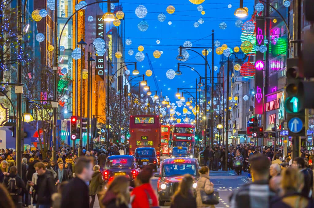 Julelys og dekorasjoner i Oxford Street, med mange mennesker som handler på romjulssalget, samt offentlig transport med busser og drosjer.