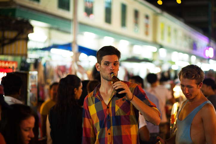 Bangkok, Thailand - March 11, 2015: Night shot of caucasian young man and tourist walking amnd drinking in Khaosan Road. Man is drinking beer. Around and behind him are many tourists and backpackers.