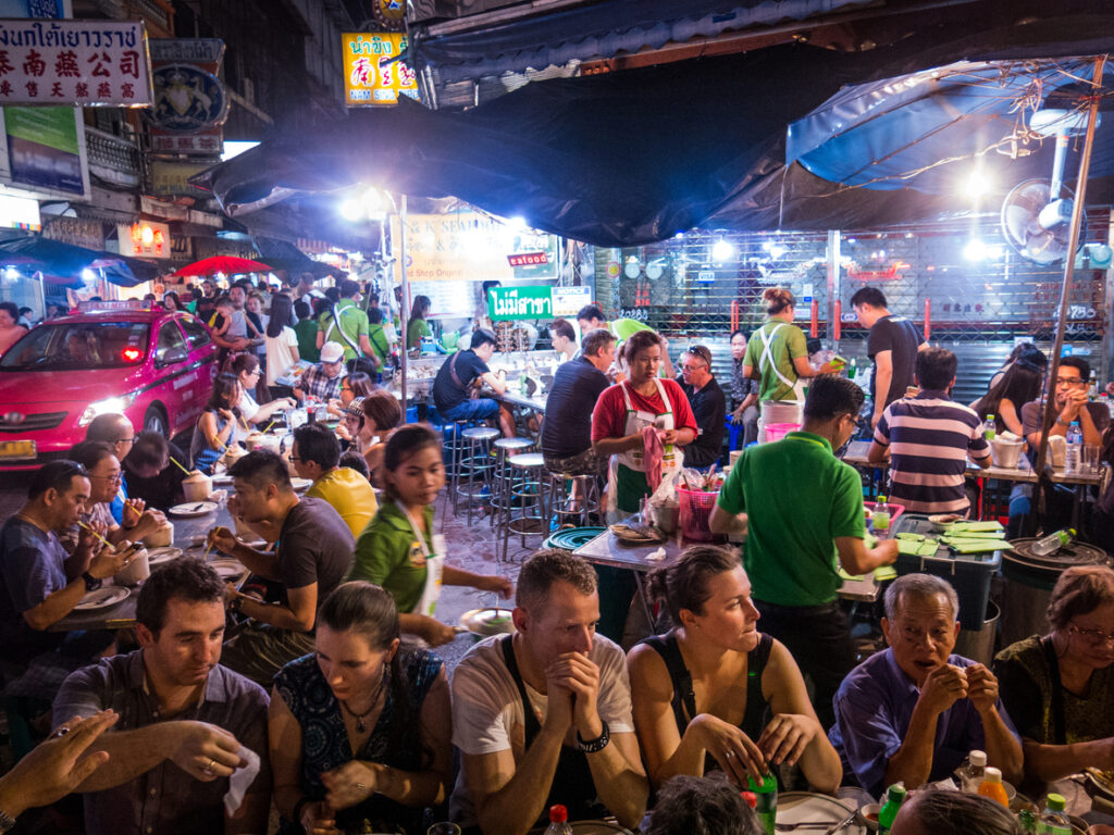 Hver kveld i uken kan du dra til den berømte Yaowarat Road i Bangkok og bli overveldet av synene, lydene og aktivitetene i dette shoppingparadiset. Neonlysene lyser opp de turistfylte gatene, markedsselgere roper til forbipasserende, og musikk dunker fra de mange barene. En av de beste tingene du kan gjøre er å ta en stol og bare se den merkelige verden gli forbi. På dette bildet spiser turistene «som lokalbefolkningen» – de omfavner reisemålet, dykker inn i kulturen og blander seg med lokalbefolkningen.