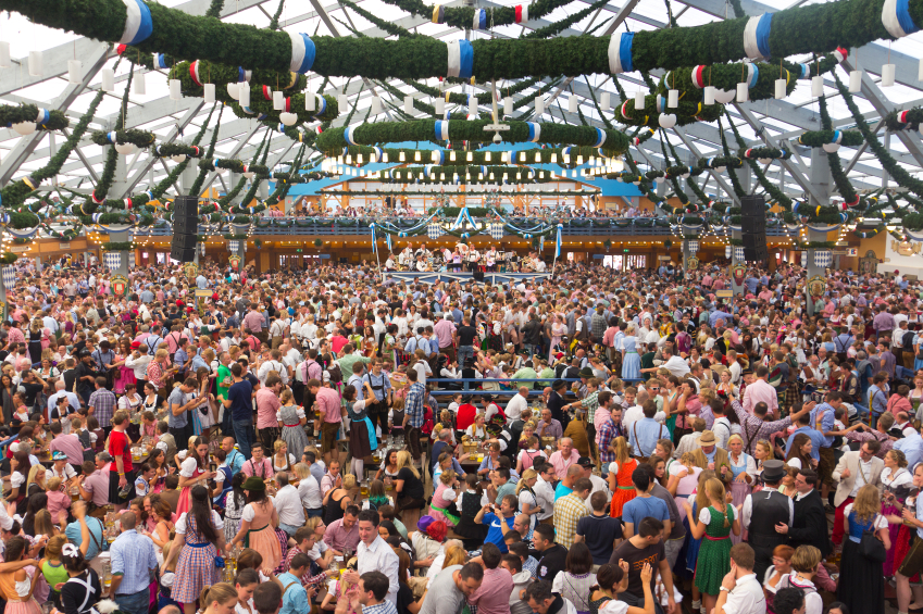 At the oktoberfest Munich. Hundreds or maybe thousands of poeple are inside one of the big beertents, sitting or standing on their benches, talking, drinking and having fun. Many of them dressed in dirndls or leather pants. In front is the band playing drinking songs.
