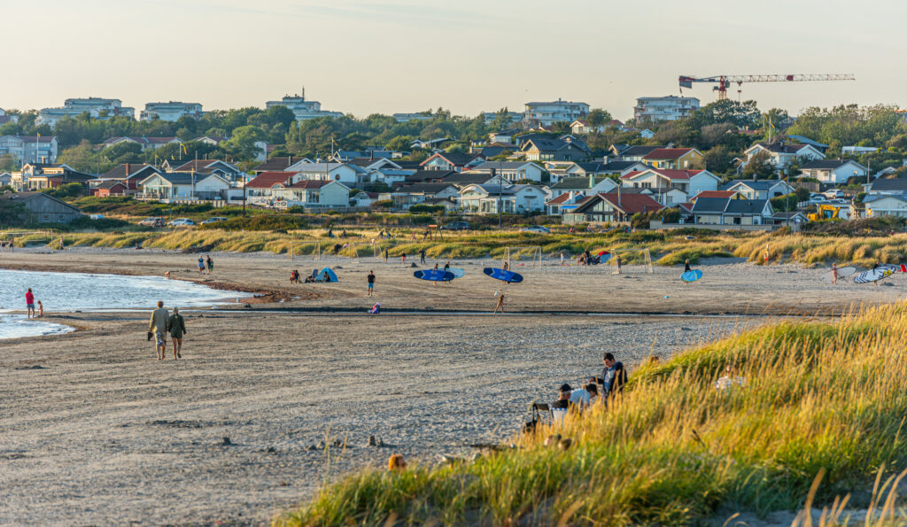 Sykkelferie langs Kattegattleden i Vaberg, på Apelviken strand. Idyllysk sandstrand med hus og folk med surfebrett. 