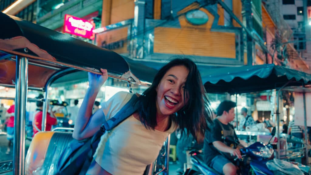 Thailandsk dame som kjører i Tuk Tuk bil, smilende og et livlig marked i bakgrunnen.