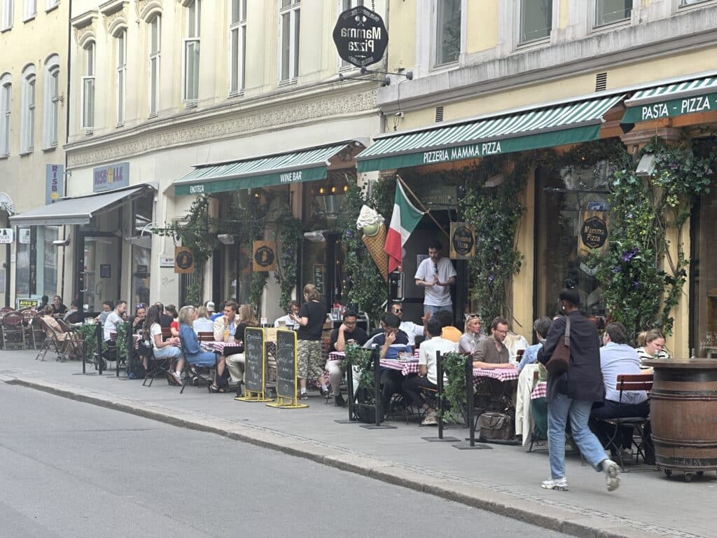 Folk som sitter utendørs restauranten og spiser pizza med italiensk flagg i bakgrunnen og mange små bord. Hva kan man gjøre i Oslo?