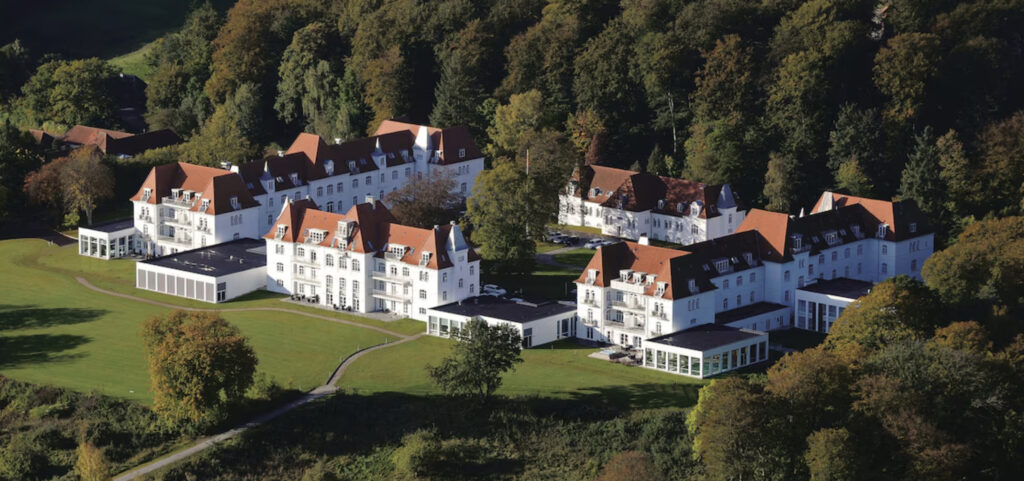 Comwell Kellers Park har store bygninger med hite vegger og brune tak. Omringet av store grønne trær.