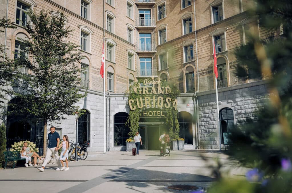 Familiehotell i Gøteborg rett ved Liseberg. Hotellinngang med stor tekst på hotellets navn og folk som kommer ut av døra. Flagg og grønne trær. 