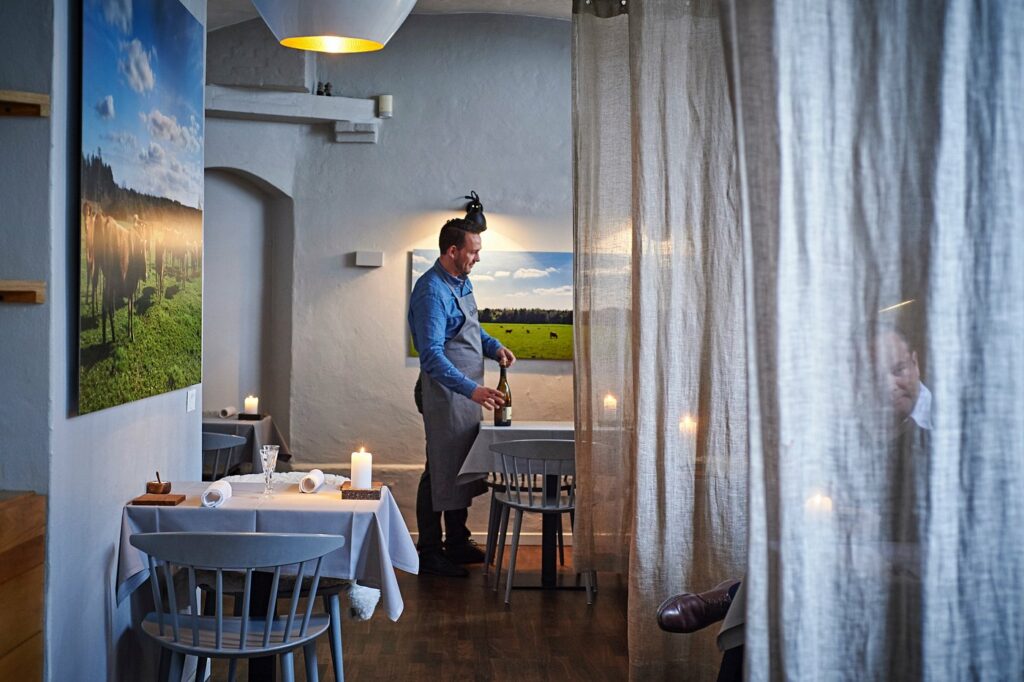 Restaurant med en mann som skal servere vin. Lun stemning med lys på bordet, stort bilde av kuer i en gård og hvite murvegger.