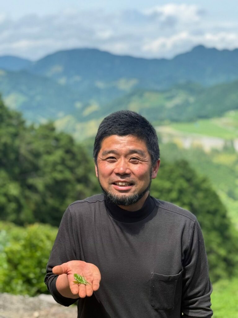 Hiroki viser frem et teblad mens han smiler til kamera. Grønt landskap i bakgrunnen.