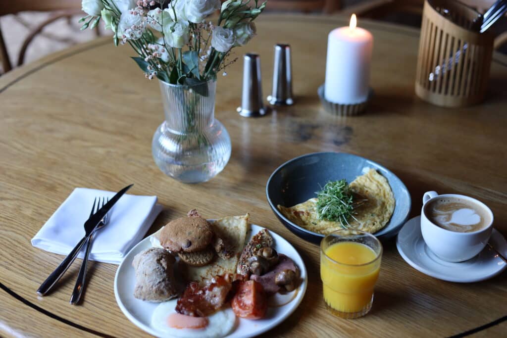 Bord med tallerkener med mat, appelsinjuice i glass og kaffekopp med latte. Bestikk, blomstervase og salt og pepper børse.