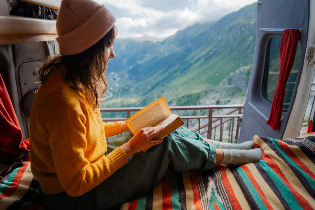 Kvinne som leser en bok i en campingbil med utsikt over Furkapasset under en reise i de sveitsiske Alpene.