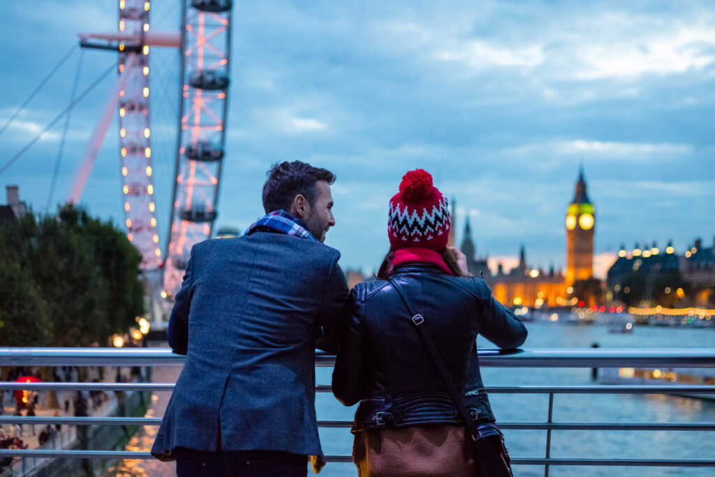Bakfra-visning av et turistpar iført vinterklær som ser mot Big Ben og London Eye om kvelden.