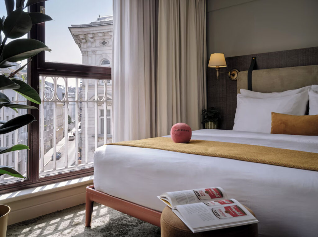 Hotell i Wien med dobbeltseng, utsikt over byen fra vinduet, bok på sittepuffen og en grønn plante. 